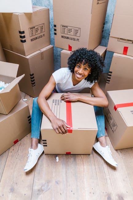 A young woman with curly light brown hair, wearing a light blue denim shirt, smiling at the camera inside a room filled with moving boxes made of plain brown cardboard, some stacked on top of each oth