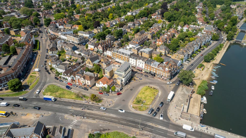 An aerial view of a residential area along Balham High Road, showing a mix of detached and terraced houses with tiled roofs, surrounded by green trees and streets with parked cars. A riverside section features a small dock with boats moored along the water's edge, adjacent to a paved walkway and a low wall. In the foreground, a section of the road is visible with moving vehicles, including cars, vans, and a delivery truck, near a small roundabout and parking areas. The image captures the urban landscape during daytime under clear weather, illustrating the typical setting for house removals and furniture transport services in Balham. [COMPANY_NAME] may utilize such locations for home relocation projects, including loading and unloading processes, packing activities, and logistical planning for local moves, as part of their removals service offering.