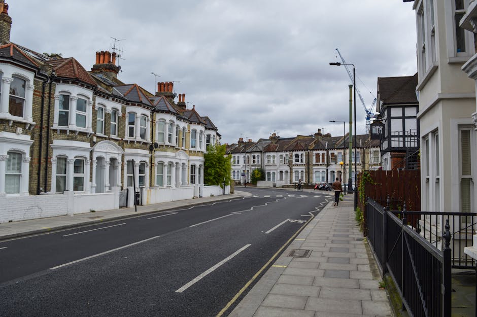 A street view of residential houses on Balham High Road, with a sidewalk running along the right side, featuring black metal railings and a series of paved slabs. The buildings are mainly terraced houses with white-painted facades and bay windows, some with decorative trim, and brickwork exteriors. On the left, there are houses with red-tiled roofs, bay windows, and chimneys, typical of Victorian architecture. The sky is overcast with grey clouds, and the street appears quiet with no visible traffic. Street lamps and a construction crane are visible in the background, indicating ongoing development. The scene captures a typical residential area prepared for home relocation or furniture transport, with a focus on the orderly environment where removals services by [COMPANY_NAME] may operate, especially during packing and loading stages of house removals and moving logistics.